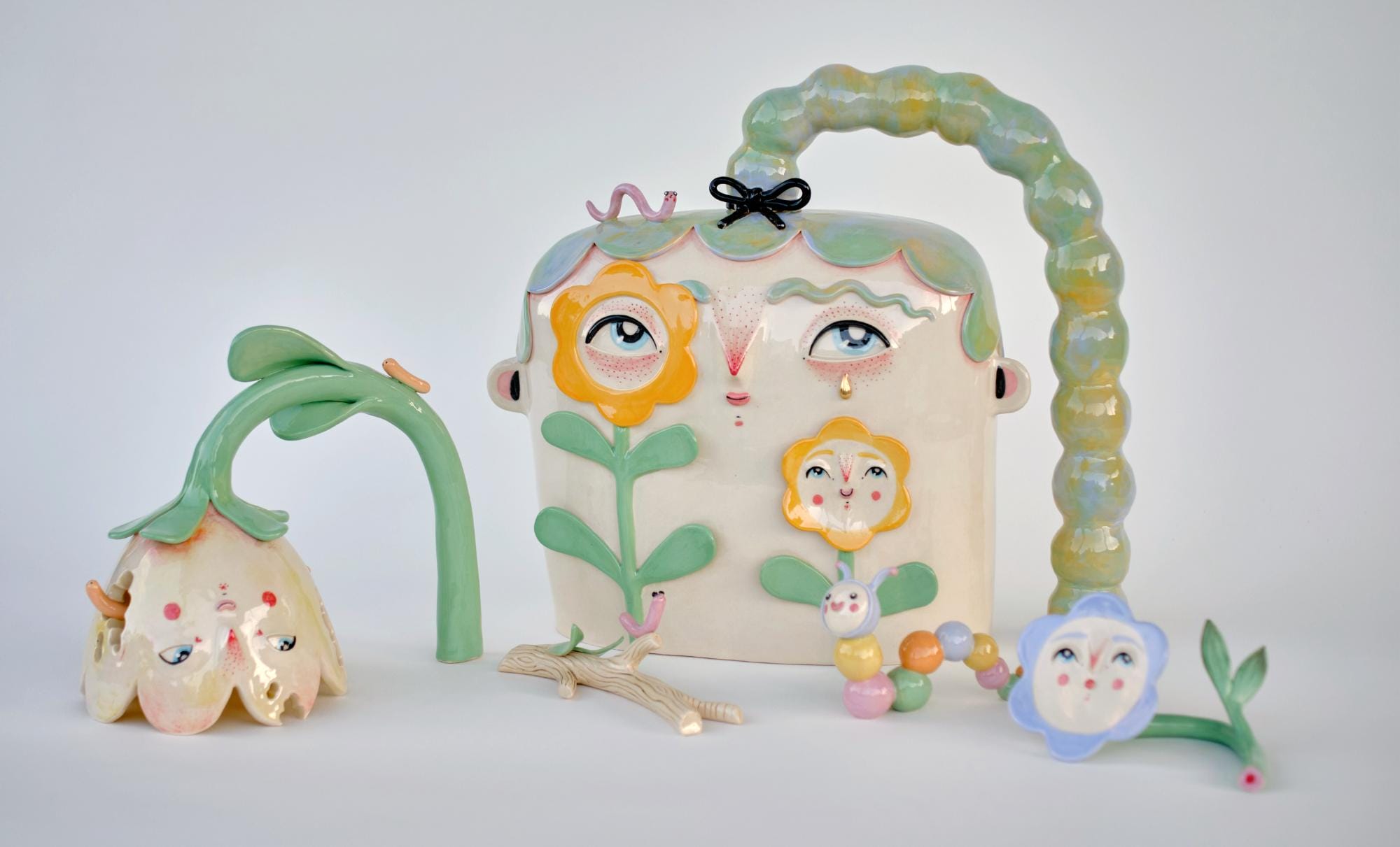 ceramic sculptures by Abi Castillo of a head with green hair and flowers with faces, surrounded by cute worms and colorful caterpillars