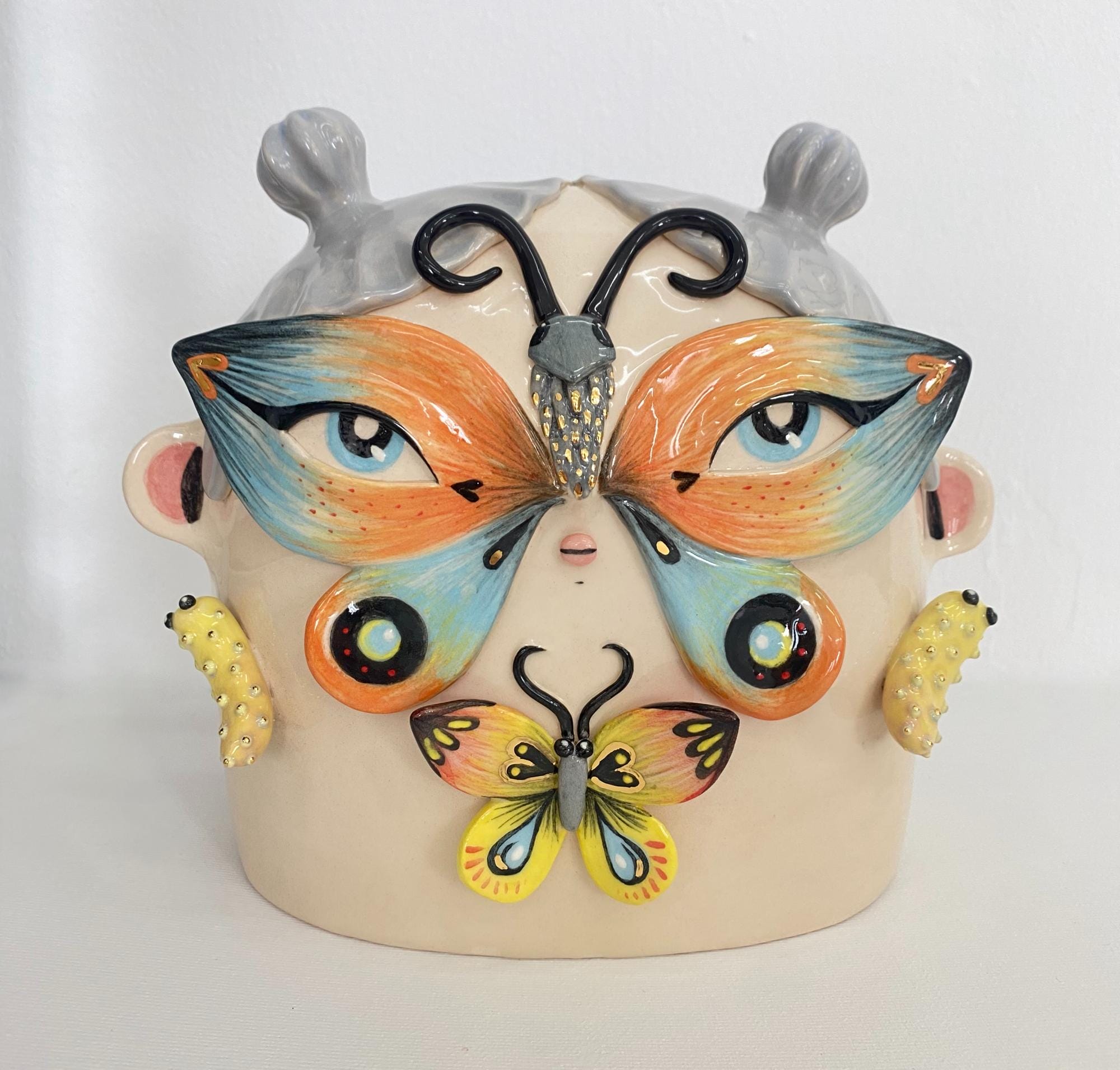 a ceramic sculpture by Abi Castillo of a head with gray hair and a masked shaped like a butterfly around her eyes, with a smaller butterfly and caterpillar details